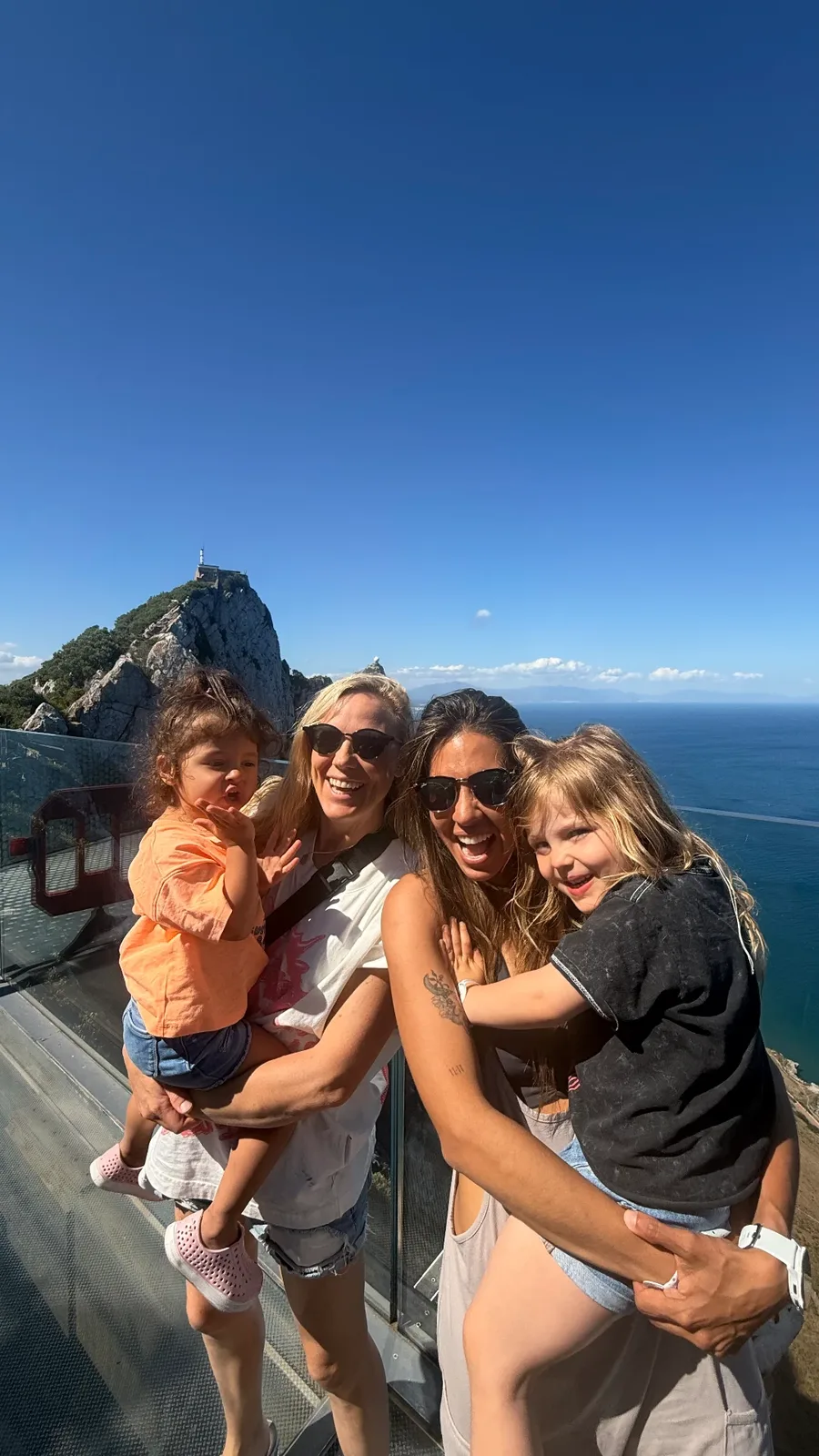 Morgan and Ale holding their two daughters on a clifftop walkway above the sea in Southern Europe