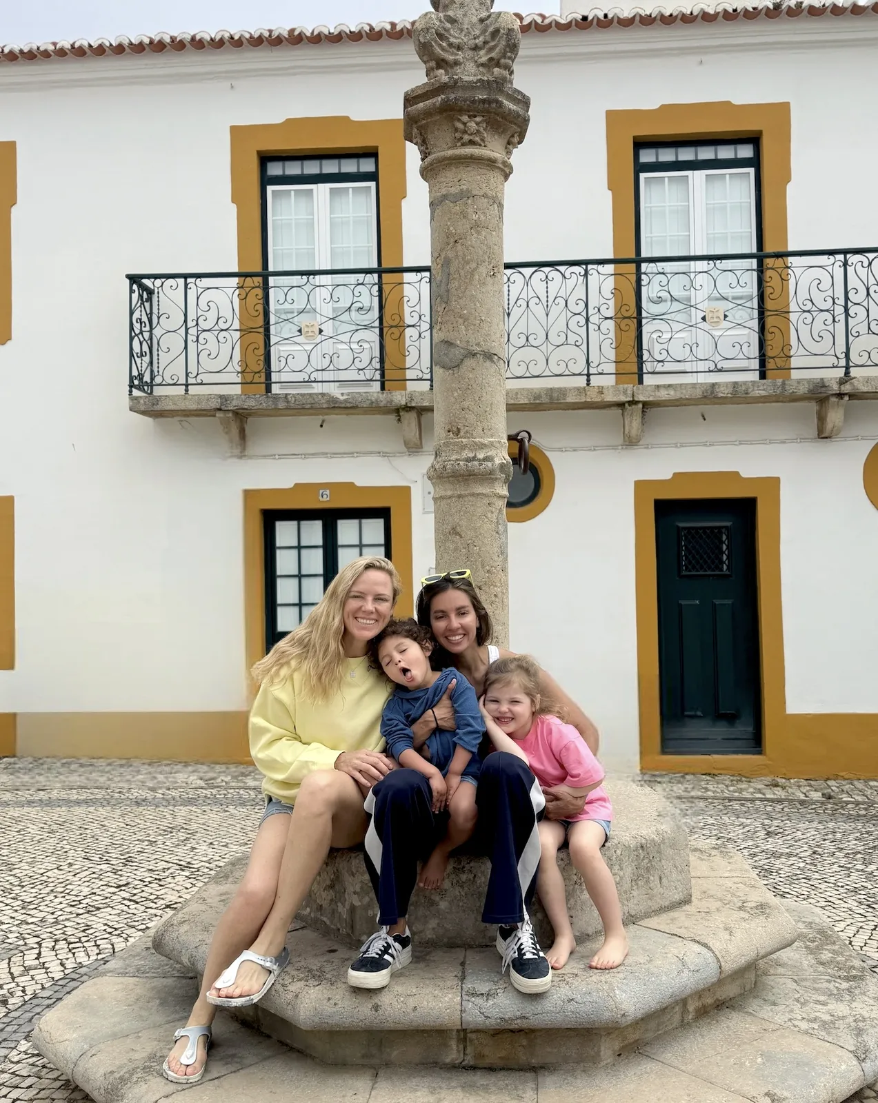 Morgan and Ale sitting with their children by a stone column in a sunlit Portuguese village square